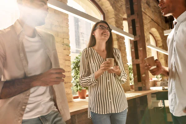Kontorsnyheter. Tre unga och glada människor i casual wear hålla kaffekoppar och diskutera något med leende när du står i det moderna kontoret — Stockfoto
