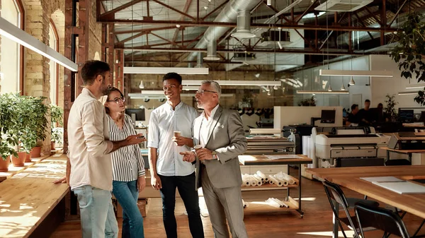 Discussie over een nieuw project. Vier collega 's die koffiebekers vasthouden en ergens over praten terwijl ze in creatief kantoor staan — Stockfoto