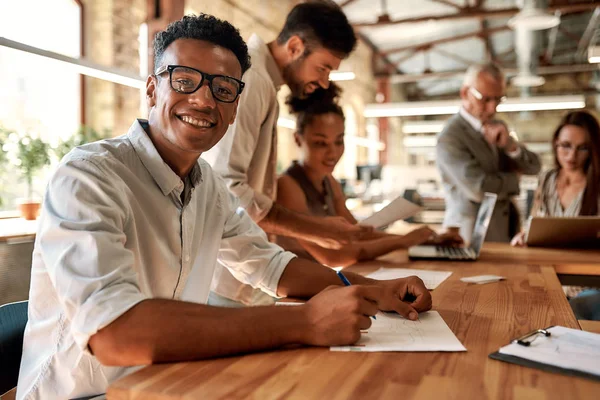 Junger, fröhlicher Afroamerikaner mit Brille, der lächelnd in die Kamera blickt, während er mit seinen Kollegen im modernen Büro arbeitet — Stockfoto