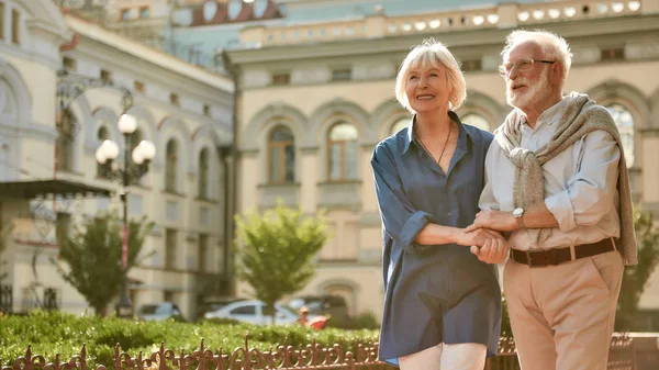 Совместное времяпрепровождение. Счастливая и красивая пожилая пара, держась за руки во время прогулки на улице — стоковое фото