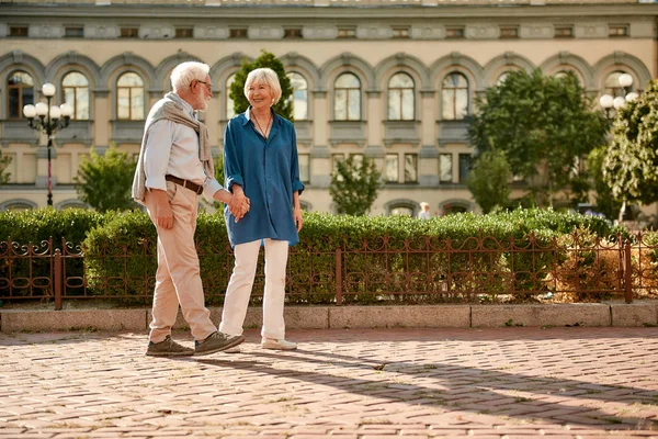 Love has no age limit. Beautiful elderly couple holding hands and looking at each other with smile while walking together outdoors