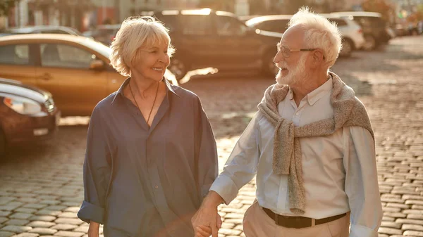 Nach Hause gehen. glückliches älteres Paar Händchen haltend und lächelnd zueinander blickend, während es gemeinsam durch die Straße geht — Stockfoto