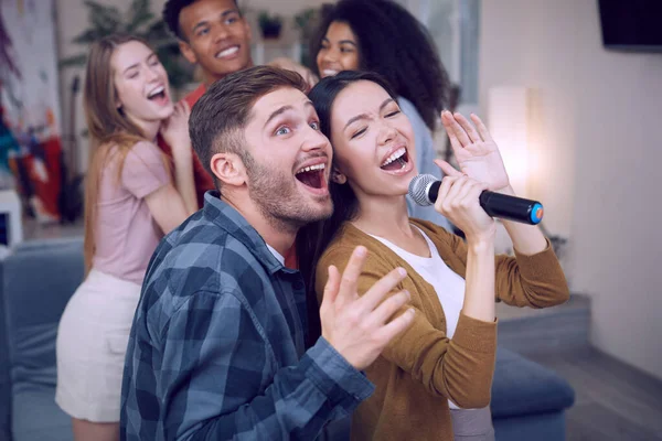 Bernyanyi duet. Pasangan muda yang bersemangat atau teman-teman memegang mikrofon dan bernyanyi bersama sambil bermain karaoke dengan teman-teman di rumah. Kelompok orang bersenang-senang, pesta karaoke — Stok Foto