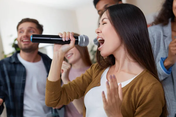 Fiesta en casa. Joven hermosa chica asiática sosteniendo micrófono y cantando mientras juega karaoke con sus mejores amigos en casa. Grupo de gente feliz divirtiéndose juntos. Enfoque selectivo — Foto de Stock