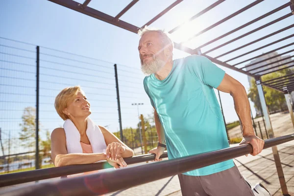 Senioren-Familie trainiert im Outdoor-Fitnessstudio. Reifer bärtiger Mann in Sportbekleidung, der Liegestütze an parallelen Barren macht. Aktives Senioren-Paar trainiert am Morgen — Stockfoto