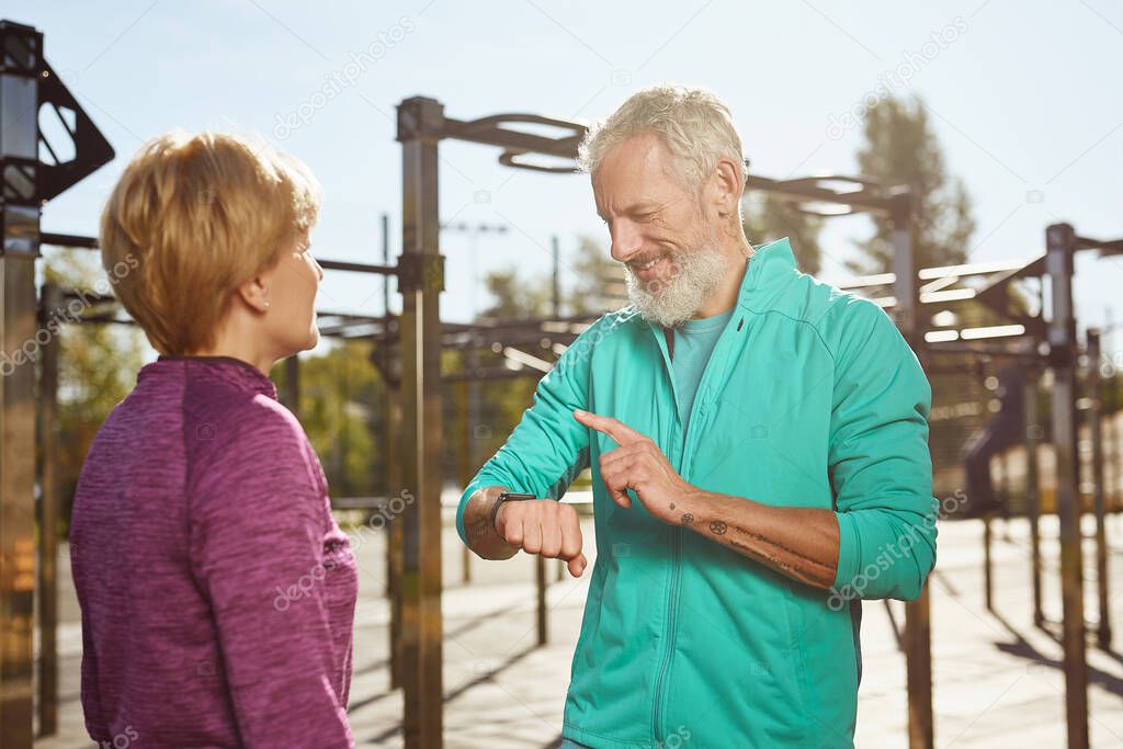 Checking fitness results. Happy senior man in sportswear looking at smartwatch and smiling while exercising with his wife at outdoor gym. Active family couple