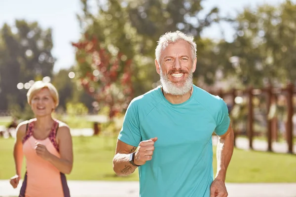 Gesund und fit bleiben. Ein glücklicher älterer Mann lächelt in die Kamera, als er am frühen Morgen gemeinsam mit seiner Frau läuft. Selektiver Fokus — Stockfoto