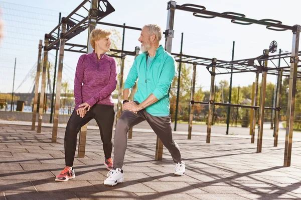 Full length of senior or mature happy couple in sportswear exercising together in the early morning at the stadium. They are looking at each other and smiling