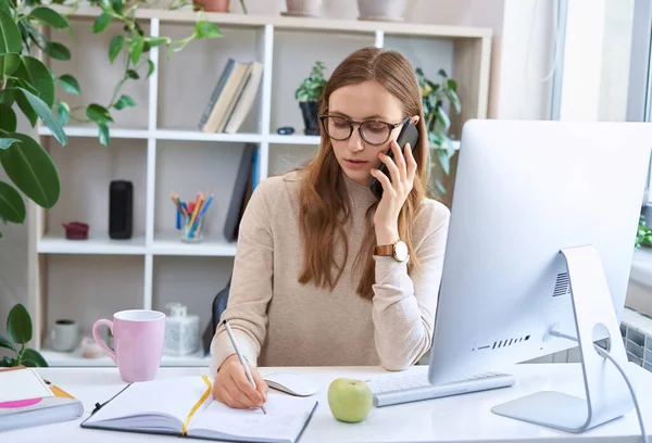 Joven mujer de negocios ocupada, atractiva empleada en gafas sentada en el escritorio, hablando por teléfono y tomando algunas notas mientras trabaja de forma remota en casa —  Fotos de Stock