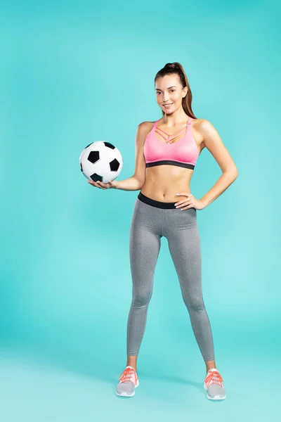 Ποδοσφαιριστής. Πλήρες μήκος της νεαρής όμορφης γυναίκας στα αθλητικά κρατώντας μπάλα ποδοσφαίρου και χαμογελώντας στην κάμερα, ενώ στέκεται πάνω από το μπλε φόντο — Φωτογραφία Αρχείου