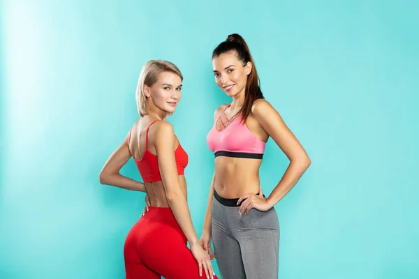 Женская сила. Две молодые и привлекательные спортивные девушки в спортивной одежде смотрят в камеру и улыбаются, стоя на синем фоне. Спорт и фитнес — стоковое фото