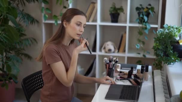 Fiatal gyönyörű lány alkalmi ruhák alkalmazó rúzs az ajkak speciális ecset, miközben nézi smink bemutató online laptop otthon. Önelkülönítés — Stock videók