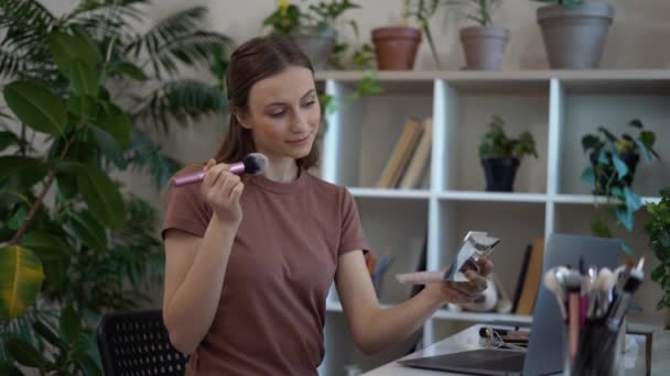 Mujer bonita joven aplicando bronceador rubor con cepillo, mirando en el espejo y sonriendo, viendo tutorial de maquillaje en línea en el ordenador portátil en casa — Vídeos de Stock