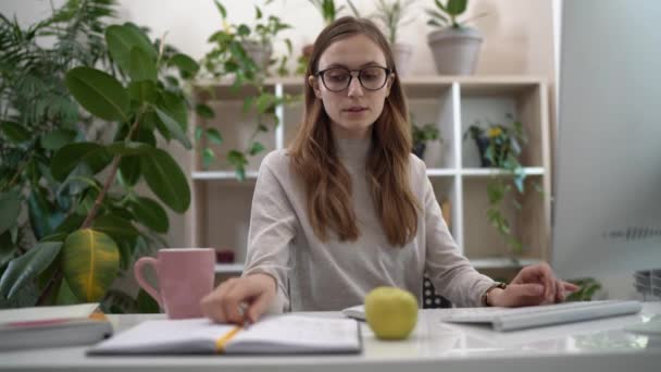 Online baan. Jonge gefocuste zakenvrouw in glazen werken vanuit huis, mooie vrouwelijke werknemer met behulp van de computer en het maken van een aantal notities in een notebook tijdens het werken thuis kantoor — Stockvideo