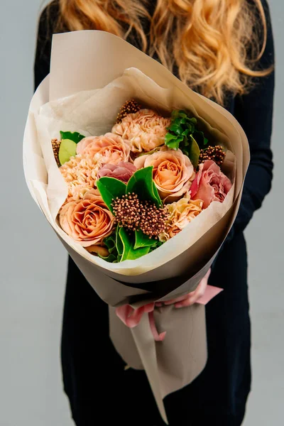 Açık arkaplandaki kızıl saçlı çiçekçinin ellerinde bej renkli bir çiçek buketi. Gül, karanfil, pittosporum, skimmia.