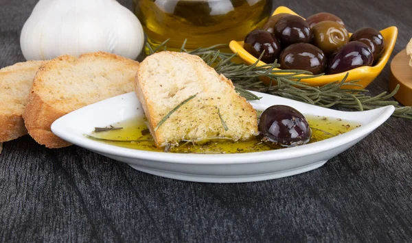 The bread dipped in olive oil with olive. Greek olive oil bread dip. Italian Bread with Oil for Dipping with Herbs & Spices. Olive oil sauce in white bowl & Greek olives on wood background.