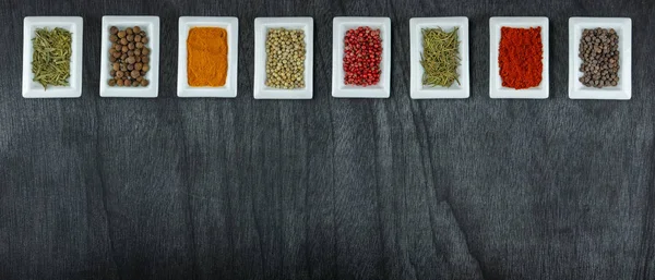 Spices and herbs on wooden table. Pepper, rosemary, tarragon, allspice, turmeric. Colorful spices in white square bowls on grey background. Seasonings for cooking. Copy space. High resolution. HD