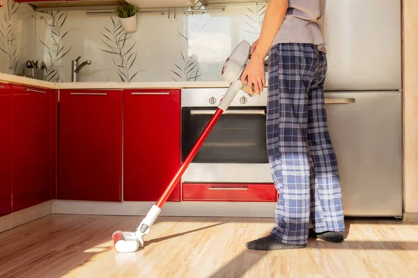 Man in home clothes cleaning house with red wireless vacuum cleaner. Red kitchen and sunshine in floor.
