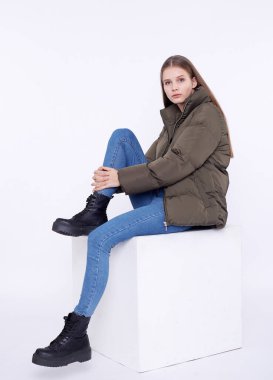 Beyaz arka planda beyaz bir kübün üzerinde oturan haki renkli ceketli genç kız..