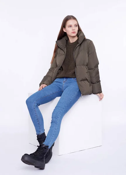 Young girl in a khaki down jacket sitting on a white cube isolated on a white background. — Stock Photo, Image