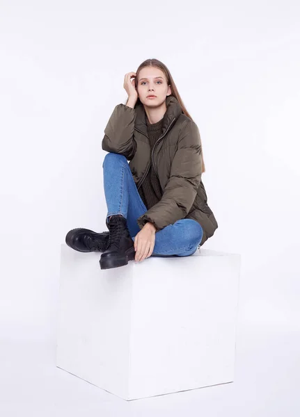 Menina jovem em um casaco cáqui sentado em um cubo branco isolado em um fundo branco . — Fotografia de Stock