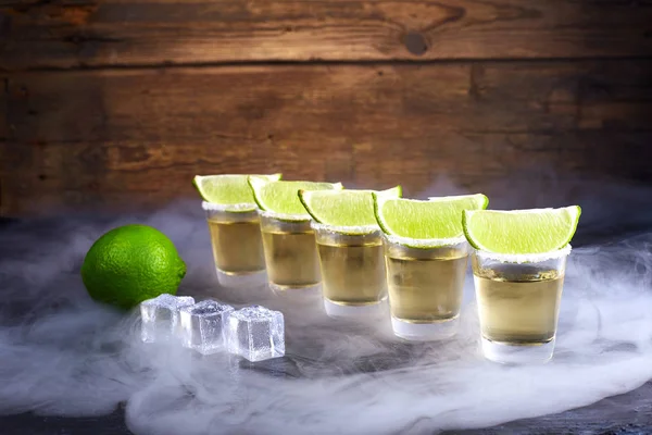 Mexican tequila gold in short glasses with salt, lime slices and ice on a wooden table. Smoke.