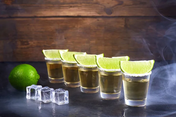 Μεξικάνικη τεκίλα σε μικρά ποτήρια με αλάτι, φέτες λάιμ και πάγο σε ξύλινο τραπέζι. Καπνός. — Φωτογραφία Αρχείου