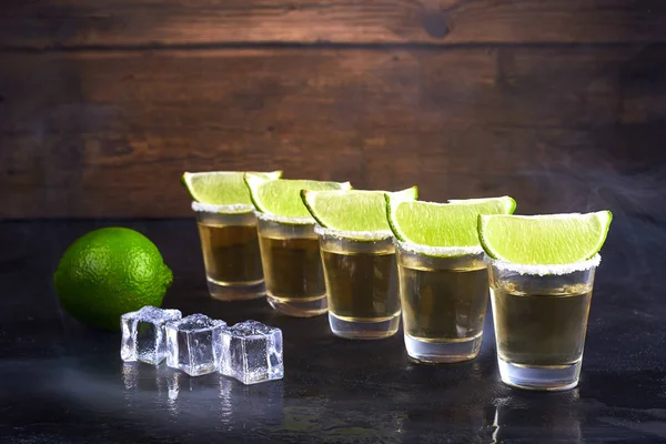 Oro de tequila mexicano en vasos cortos con sal, rodajas de lima y hielo sobre una mesa de madera. Humo. . — Foto de Stock