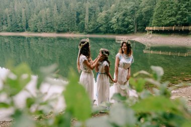 Anne ve iki kızı beyaz elbiseler kır çiçekleri çelenk ile göle yakın yürüyüş