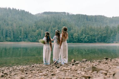 Anne ve iki kızı beyaz elbiseler kır çiçekleri çelenk ile göle yakın yürüyüş