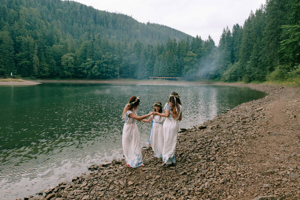 Mom and two daughters walking near the lake in white dresses with wreaths of wildflowers