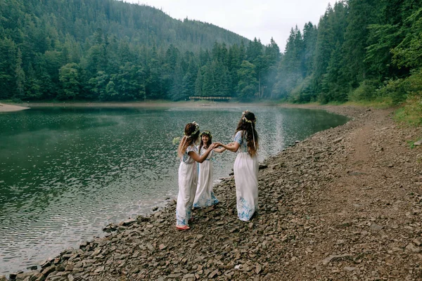Mom and two daughters walking near the lake in white dresses with wreaths of wildflowers