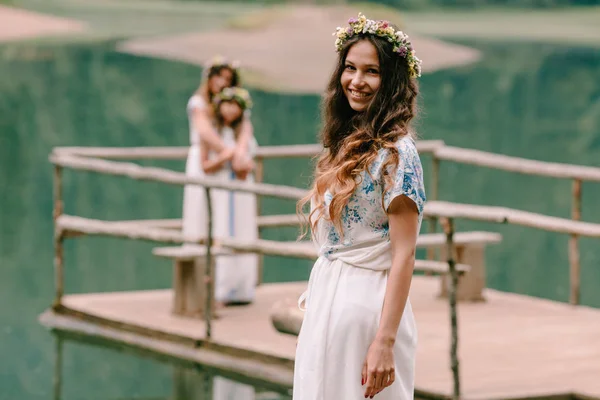 Anne ve iki kızı beyaz elbiseler kır çiçekleri çelenk ile göle yakın yürüyüş