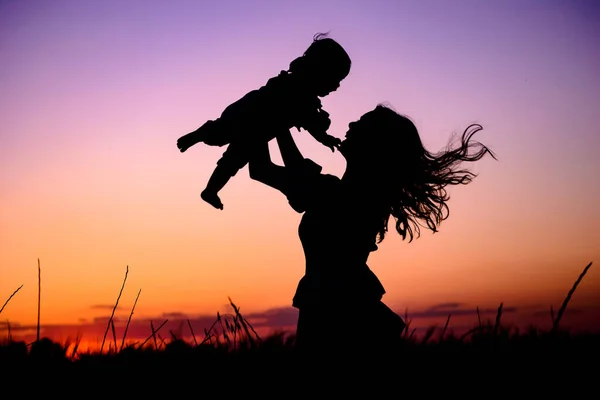 Silhouette happy mother and baby. Mother plays with her baby in her arms under the rays of the sunset in a meadow. Motherhood concept. Happiness, inspiring, joyful moments. Mother\'s day concept.