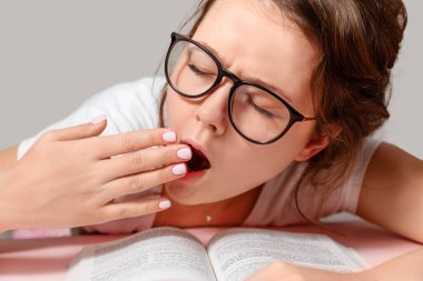 Gözlüklü genç bir kadın kitap okumaktan yoruldu. Esniyor. Evde eğitim.