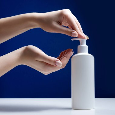 Beyaz bir masada beyaz bir şişe dezenfektan. Kız ellerini antibakteriyel jelle tedavi etmek için makineye basıyor. Virüs tasarımı konsepti. Minimalizm.