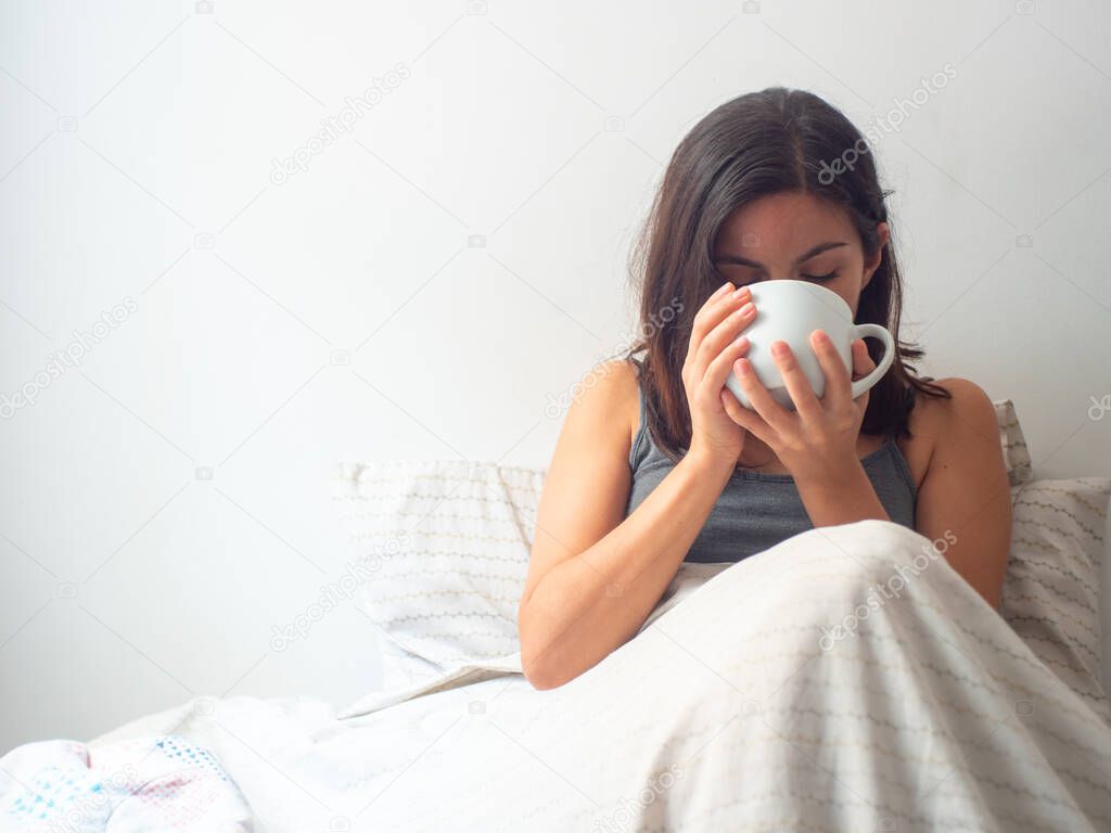 Young Brown Hair Hispanic Female Drinks Tea or Coffee in a White Cup in her Bed in Quarantine