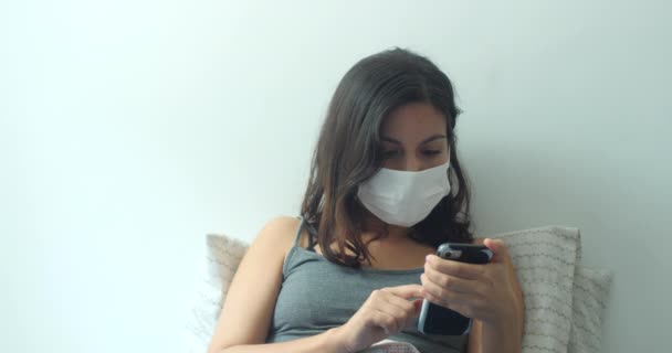 ヒスパニック系の若い茶色の髪の女性は インフルエンザ ウイルス ウイルス保護のための使い捨ての顔マスクを着用してください 彼女は家にいて隔離のためにベッドで電話をかけてる — ストック動画