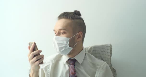 一个白种人年轻的金发男子穿着白衬衫 头戴领带 戴着可处置的口罩 为咳嗽 病毒和病毒提供保护 他待在家里 在床上喊着要他的手机 — 图库视频影像