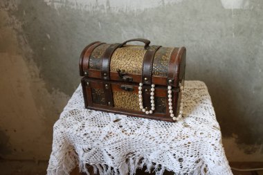 Closed dusty beautiful brown chest on a white knitted tablecloth against the concrete gray streaked wall. Two strings of pearls hang from the chest. clipart