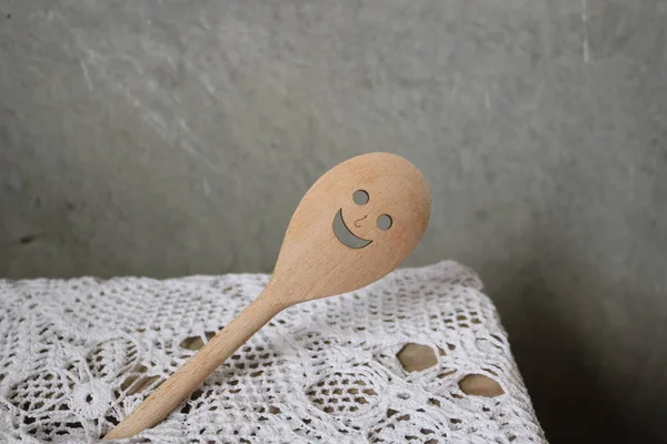A smiling wooden spoon with holes for mouth and eyes against a white lace tablecloth and a gray wall. Spoon-skimmer wants a good appetite and a slim figure. With its help, you can easily get the best the best.Life is beautiful under any circumstances