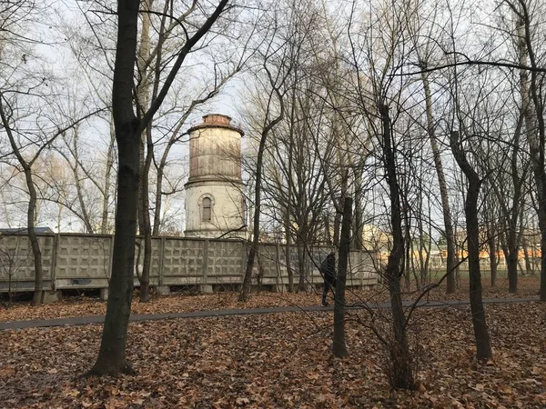 An old water tower behind an old fence among bare trees and fallen leaves against a gray autumn sky. By on the path behind the trees is a man. Photos from a mobile phone during a fall sunset.