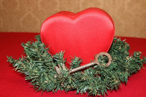 A red satin heart in green artificial fir branches with an old antique large key on a Red table against the beige wall.