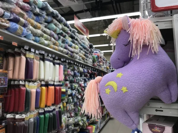 A toy plush lilac pony with a pink mane and yellow stars hangs against the background of shelves with goods for needlework. Mobile photo with store lighting.