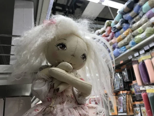 A homemade rag doll in pastel colors hangs on the background of shelves with goods for needlework. Mobile photo with store lighting.