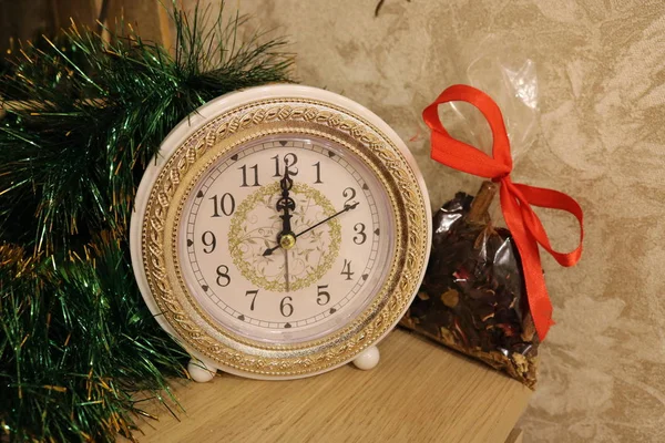 A white and gold vintage clock stands on a wooden table against a background of green tinsel and a transparent bag of spices.