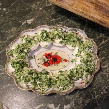 Yeşil sebzelerle doldurulmuş yumurtalar ve ortasında çiçek deseni olan oval beyaz bir tabak yeşil bir taş masanın üzerinde duruyor..