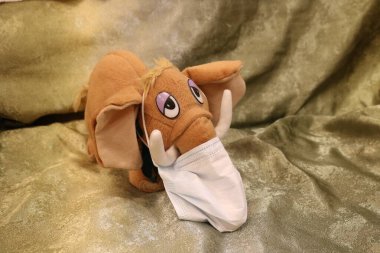 Doldurulmuş oyuncak bir mamutun üzerinde beyaz bir tıbbi maske. Sağdaki yeşil kumaşın arka planına bakıyor.. 