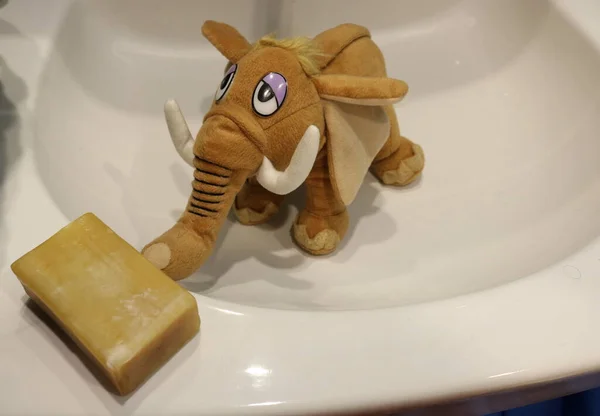 A toy stuffed mammoth standing in the sink touches a large piece of simple household soap lying on the edge of the sink with its trunk. The photo was taken under home lighting.