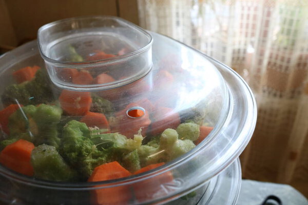 A fragment of a transparent plastic steamer with green and orange vegetables lying in it against the background of the kitchen. It was photographed in room light. 